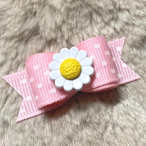 May include: A pink hair bow with white polka dots and a white daisy with a yellow center. The bow is made of ribbon and has a classic bow shape. The daisy is a decorative embellishment.