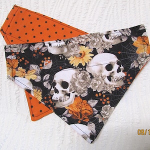 May include: Two Halloween-themed dog bandanas. One is orange with black polka dots. The other is black with white skulls, flowers, and spiderwebs. The bandanas are made of fabric and are likely handmade.