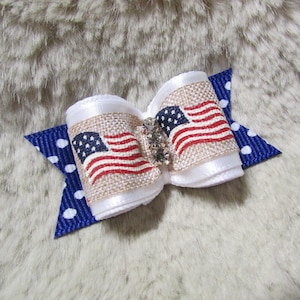 May include: A small, white and blue bow with a burlap center featuring a printed American flag design. The bow has a blue and white polka dot accent on the back.