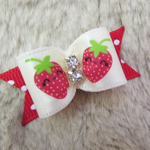 May include: A white satin bow with a red polka dot accent and two smiling strawberry graphics. The bow features two clear crystal embellishments in the center. The strawberries have green leaves and are smiling.