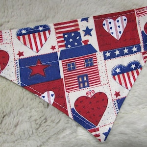 May include: Patriotic dog bandana featuring a patchwork design with red, white, and blue elements. The bandana showcases hearts, stars, and house motifs, all set against a cream-colored background. The fabric is stitched with white thread.
