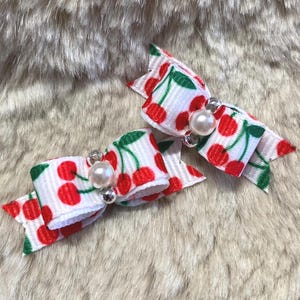 May include: Two white ribbon bows with a red cherry pattern and green stems. Each bow has a pearl and silver bead embellishment in the center. The bows are on a soft, textured background.