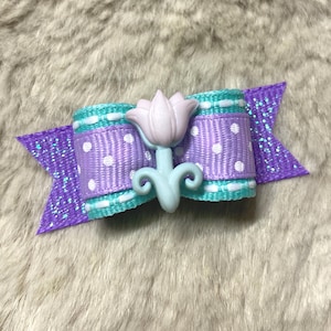 May include: A purple and turquoise polka dot bow with a light blue flower accent. The bow is made of ribbon and has a glitter finish.