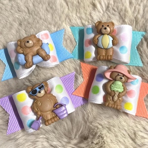 May include: Four hair bows featuring brown teddy bear figures. One bear floats on a blue ring, one wears a yellow vest, one wears blue sunglasses with a purple bucket, and one wears a pink hat holding an ice cream cone. The bows have white ribbon with colorful polka dots and solid colored ribbon edges.