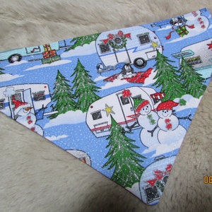 May include: A blue and white dog bandana with a repeating pattern of snowmen, Christmas trees, and camper trailers. The bandana has a festive holiday theme with red and green accents, perfect for a pet.