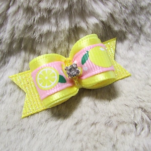 May include: A yellow and pink bow with a lemon print and a rhinestone in the center. The bow is on a light brown fuzzy surface.