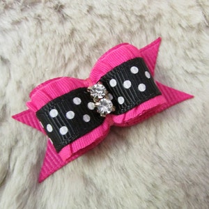 May include: A bright pink bow with a black ribbon center featuring white polka dots. The bow is adorned with two clear, sparkling rhinestones. The background is a soft, textured, light gray surface.