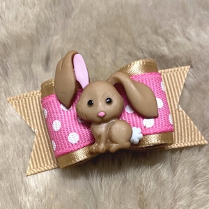 May include: A decorative hair bow featuring a brown bunny figurine with pink inner ears, set on a pink ribbon with white polka dots. The bow has a gold ribbon base, perfect for Easter or spring.