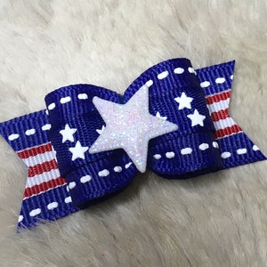 May include: A blue and red bow with white stars and a white glitter star in the center. The bow is made of ribbon and is perfect for adding a touch of patriotism to any outfit.