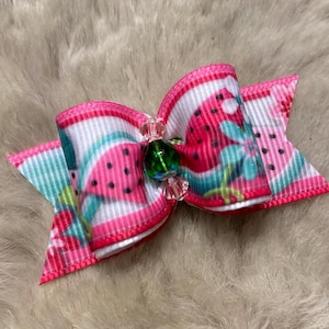 May include: A small, decorative bow featuring a watermelon pattern in pink, white, and teal. The bow is embellished with a green and pink jewel in the center. The bow is likely for a pet or hair accessory.