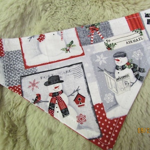 May include: A festive dog bandana featuring a winter holiday theme. The fabric is patterned with snowmen, snowflakes, and postcard designs in red, white, and gray. The bandana has a red polka dot border and includes the text "AIR MAIL".