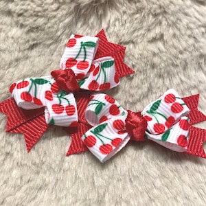 May include: Two red and white cherry print hair bows. The bows have a red ribbon base with a starburst design. The bows are white with a repeating pattern of red cherries and green stems.