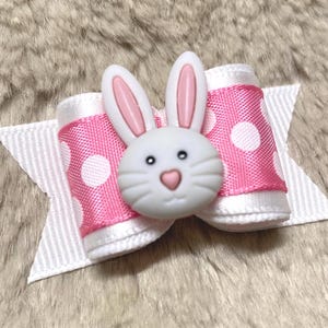 Peut inclure: Un nœud décoratif avec un visage de lapin blanc et un ruban rose à pois. Le lapin a des oreilles roses et un nez en forme de cœur. Le nœud est posé sur un ruban blanc.