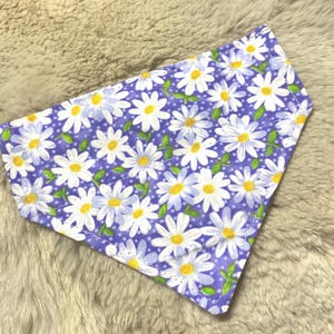 May include: A triangular pet bandana featuring a daisy pattern. The bandana has a light purple background with white daisies, yellow centers, and green leaves. The edges are neatly stitched with green thread.