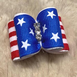 May include: A patriotic bow featuring a blue center with white stars and red and white striped sides. The bow is embellished with a row of clear rhinestones. This festive accessory is perfect for pets.