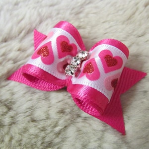 May include: A pink and white bow with a glittery heart pattern and a rhinestone embellishment. The bow is made of satin ribbon and is perfect for adding a touch of sparkle to any outfit.