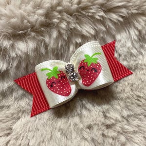 May include: A hair bow constructed from white ribbon featuring a repeating pattern of smiling red strawberries with green leaves, layered over a solid red ribbed ribbon base, with two small clear rhinestones centered on the front of the bow.
