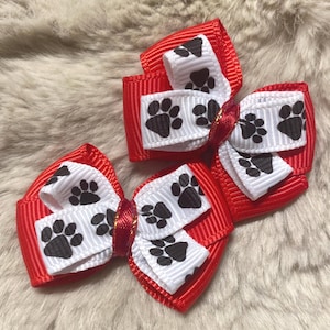 May include: Three red and white dog bow ties with black paw prints. The bows are made of grosgrain ribbon and are arranged on a fluffy, light brown surface. The bows are likely for a pet.