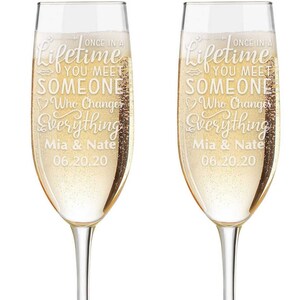 Puede incluir: Dos copas de champán transparentes llenas de líquido dorado. Las copas están grabadas con el texto "Once in a lifetime you meet someone who changes everything" y los nombres "Mia & Nate" y la fecha "06.20.20".