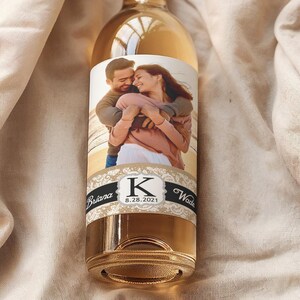 Pode incluir: Uma garrafa de vinho branco de vidro transparente com um r&oacute;tulo personalizado que mostra uma foto de um casal se abra&ccedil;ando. O r&oacute;tulo tem um design preto e dourado com a letra "K" no centro. A garrafa est&aacute; sobre uma superf&iacute;cie de tecido branco.