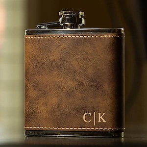 May include: A brown leather flask with a stainless steel cap and trim. The flask has a stitched border and the initials "C|K" are visible on the front. The flask is rectangular and appears to be empty.