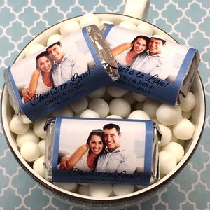 May include: Three small chocolate bars wrapped in silver foil with personalized labels. The labels feature a photo of a couple and the text "Cheers to Love!" with the couple's names and wedding date.