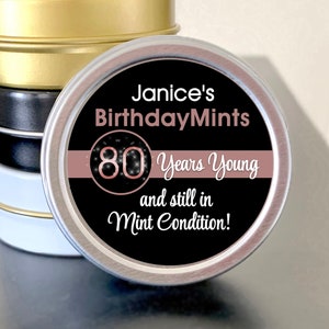 May include: A stack of four small, round tins with gold, black, and white lids. The front tin has a label that reads "Janice's Birthday Mints" with "80 Years Young and still in Mint Condition!" in white and rose gold.