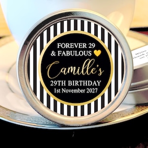 May include: A round, silver tin with a black and white striped background. The center features a black circle with gold text that reads "FOREVER 29 & FABULOUS Camille's 29TH BIRTHDAY 1st November 2027". A gold heart is also present.