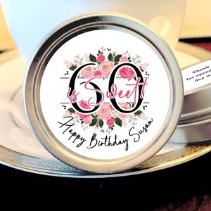 May include: A round silver tin with a white label featuring pink flowers and the text "60 is Sweet!" with a black and white striped background. The label also includes the text "Happy Birthday Susan".
