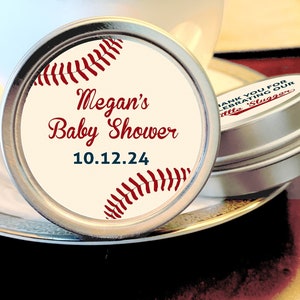 May include: A round, silver tin with a white label featuring a red baseball graphic and the text "Megan's Baby Shower 10.12.24".