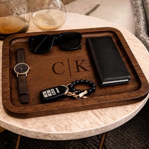 May include: A rectangular dark wood valet tray featuring the engraved letters C and K separated by a vertical line. The tray holds a black wristwatch, black sunglasses, a black wallet, a car key fob, and a black beaded bracelet with a gold charm.