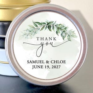 May include: Stack of small, round metal tins in gold, black, and white. One tin is in focus, featuring a white label with green foliage and the words "Thank you" and the names "Samuel & Chloe" and the date "June 19, 2027".