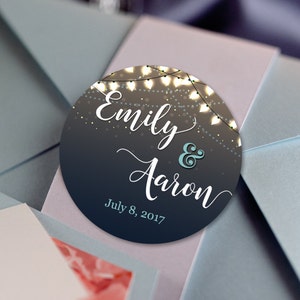 May include: A blue envelope with a white and light blue circle sticker. The sticker has the text "Emily & Aaron" in white cursive font and "July 8, 2017" in black font. The sticker has a string of lights around the edge.