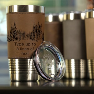 May include: A stainless steel travel mug with a brown leatherette wrap and a clear lid. The mug has a black silhouette of a forest scene and the text "Type up to 3 lines of text".