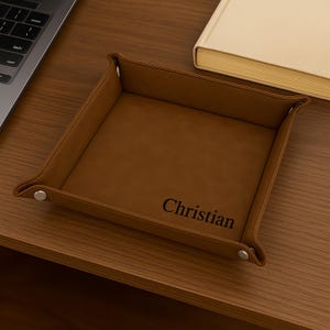 Pode incluir: Uma bandeja de couro castanho com o nome "Christian" gravado em preto. A bandeja tem formato quadrado com bordas elevadas e &eacute; fixada com bot&otilde;es de press&atilde;o prateados. A bandeja est&aacute; sobre uma superf&iacute;cie de madeira.