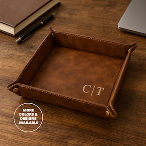 May include: A brown leather valet tray with stitched edges and snap closures. The tray features the initials "C/T" in the center. The image also includes a pen, notebook, and laptop on a wooden surface. The text "MORE COLORS & DESIGNS AVAILABLE" is in a white circle.