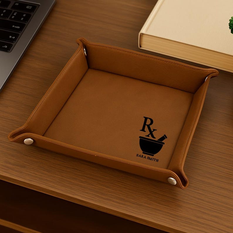 Pode incluir: Uma bandeja de couro castanho com formato quadrado e bordas elevadas. Apresenta um gr&aacute;fico preto de almofariz e pil&atilde;o com as letras "Rx" e o nome "SARA SMITH" impressos no interior. A bandeja est&aacute; sobre uma superf&iacute;cie de madeira.