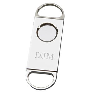May include: A silver cigar cutter with a circular blade and a rectangular body. The cutter has a loop at the top and bottom for easy handling. The initials "DJM" are engraved on the front.