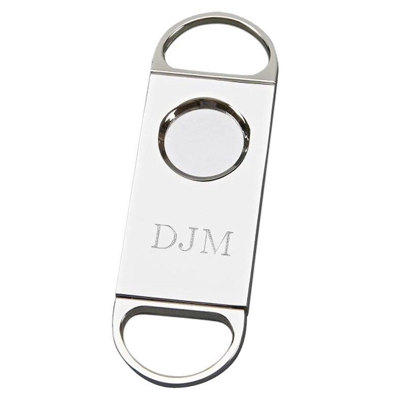 May include: A silver cigar cutter with a circular blade and a rectangular body. The cutter has a loop at the top and bottom for easy handling. The initials "DJM" are engraved on the front.