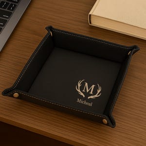 Pode incluir: Bandeja de valet em couro preto com um design personalizado. A bandeja apresenta uma borda costurada, rebites dourados e um design personalizado com a letra "M" e o nome "Micheal".