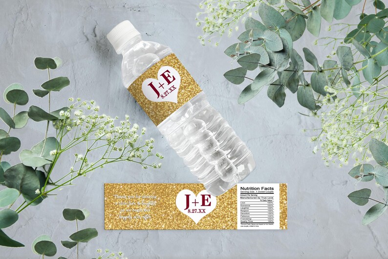 Op de afbeelding: Gouden glitter bruiloft waterfles label met een wit hart en de tekst "J+E 8.27.XX". Het label bevat ook de tekst "Bedankt dat je met ons feestviert!" en een voedingswaardelabel.