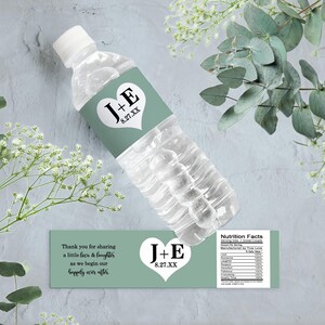 Op de afbeelding: Een groen en wit bruiloftwaterfleslabel met de tekst "J+E 8.27.XX" en een hartontwerp. Het label bevat ook de tekst "Bedankt voor het delen van een beetje liefde en gelach terwijl we beginnen aan ons geluk voor altijd."