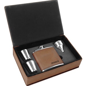 May include: A brown leather-wrapped flask with a silver top and two silver shot glasses. The flask is engraved with the name "JOHN". The flask and shot glasses are in a brown leather box with a black interior.