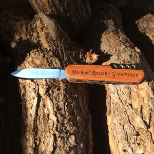 May include: A brown wooden pocket knife with a silver blade. The knife has a personalized inscription that reads "Michael Rossen - Groomsman".