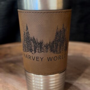 May include: A stainless steel travel mug with a brown leather wrap. The wrap features a black silhouette of a forest with the text "HARVEY WORLD" at the bottom.