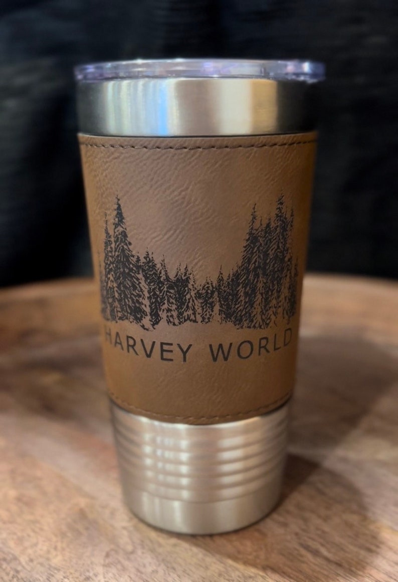 May include: A stainless steel travel mug with a brown leather wrap. The wrap features a black silhouette of a forest with the text "HARVEY WORLD" at the bottom.