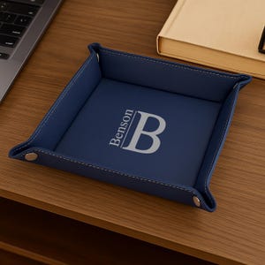 Pode incluir: Uma bandeja de couro azul marinho com uma borda costurada. A bandeja apresenta o nome "Benson" e a letra "B" em fonte prateada. A bandeja est&aacute; sobre uma superf&iacute;cie de madeira.