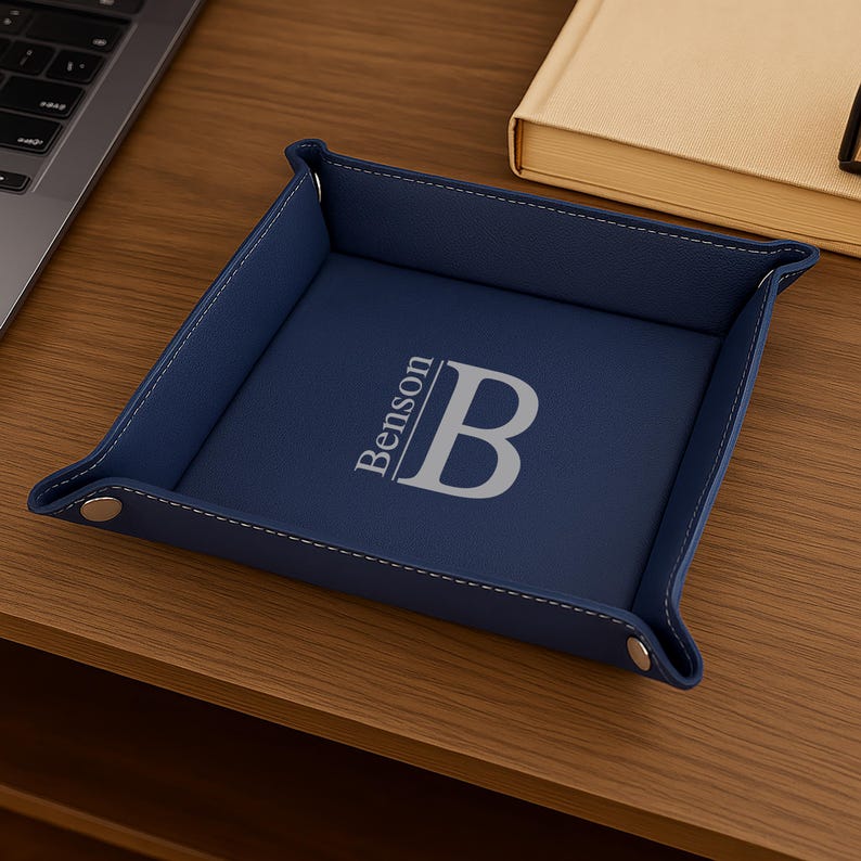 Pode incluir: Uma bandeja de couro azul marinho com uma borda costurada. A bandeja apresenta o nome "Benson" e a letra "B" em fonte prateada. A bandeja est&aacute; sobre uma superf&iacute;cie de madeira.