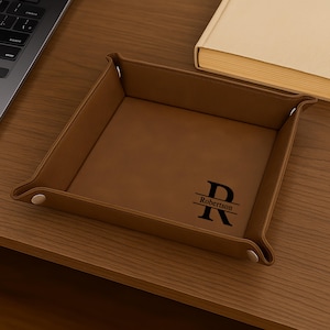 Pode incluir: Uma bandeja de couro castanho com um monograma personalizado e o nome "Robertson" em preto. A bandeja &eacute; quadrada com cantos arredondados e fechos de press&atilde;o. Um laptop e um livro est&atilde;o no fundo.