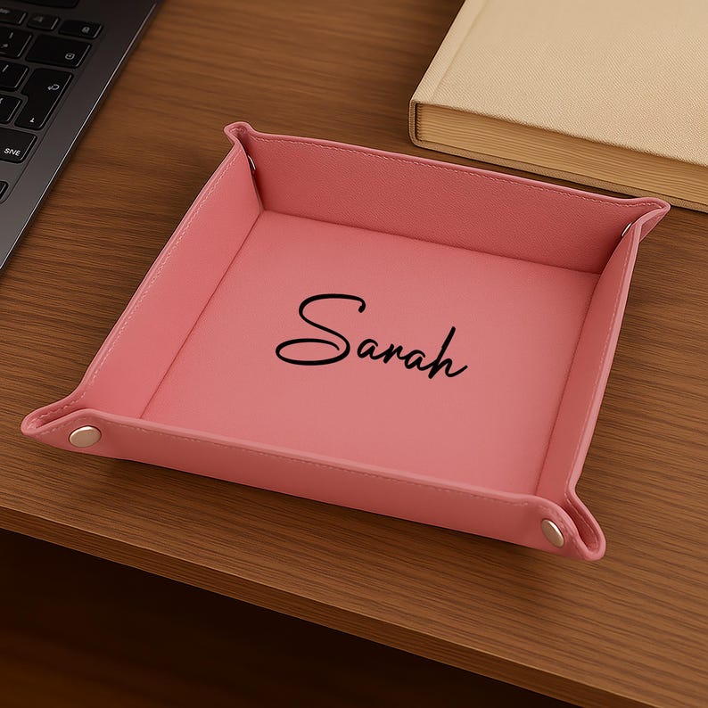 Pode incluir: Uma bandeja rosa de couro com o nome "Sarah" em escrita preta. A bandeja tem formato quadrado com cantos dobrados e fechos de press&atilde;o prateados. Um laptop e um livro est&atilde;o ao fundo.